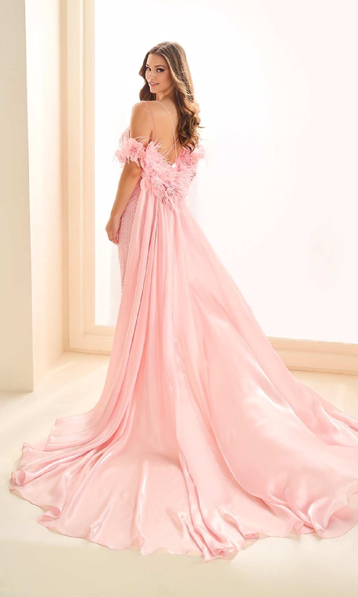 A woman stands in a brightly lit room, looking back over her shoulder, wearing the Ellie Wilde EW36027 long prom dress—pink with off-the-shoulder feather details and a detachable floor-length cape.